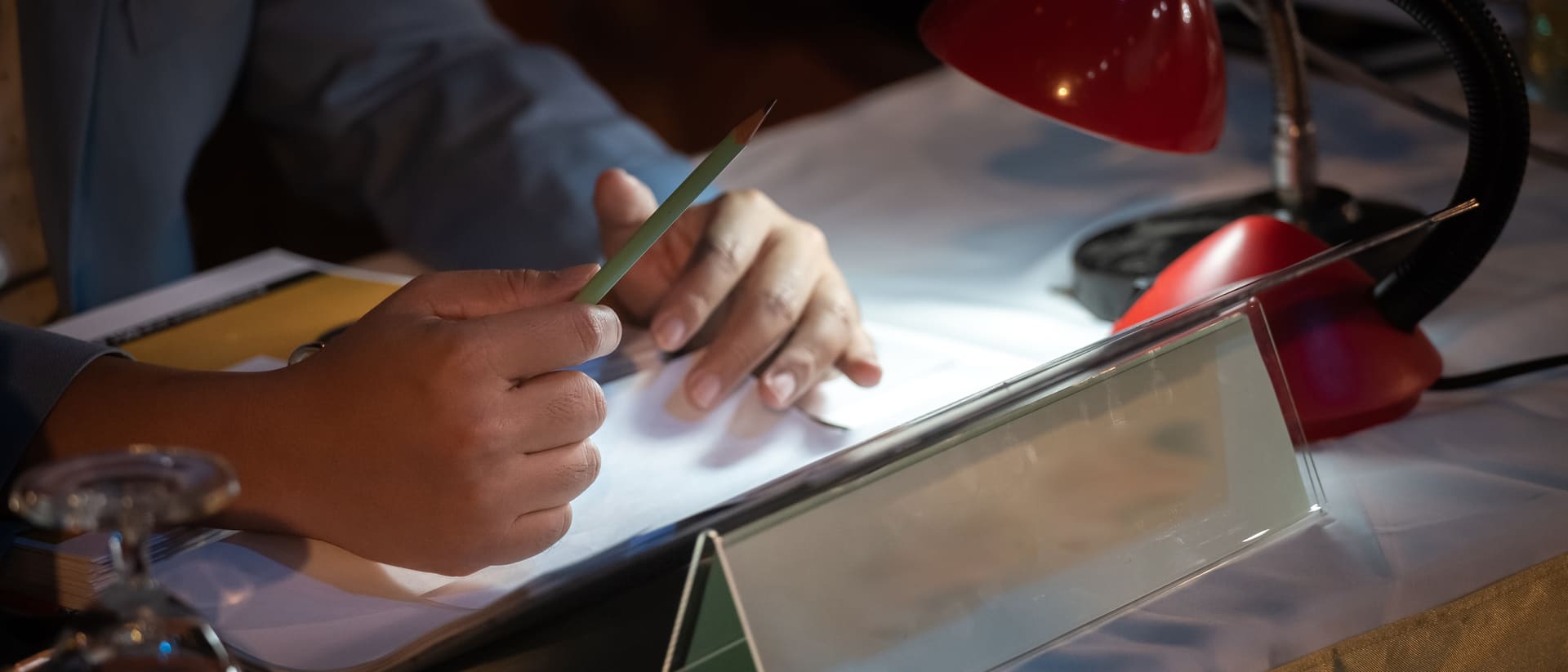 Une personne tenant un crayon, réfléchissant devant un cahier, sous une lampe de bureau rouge.