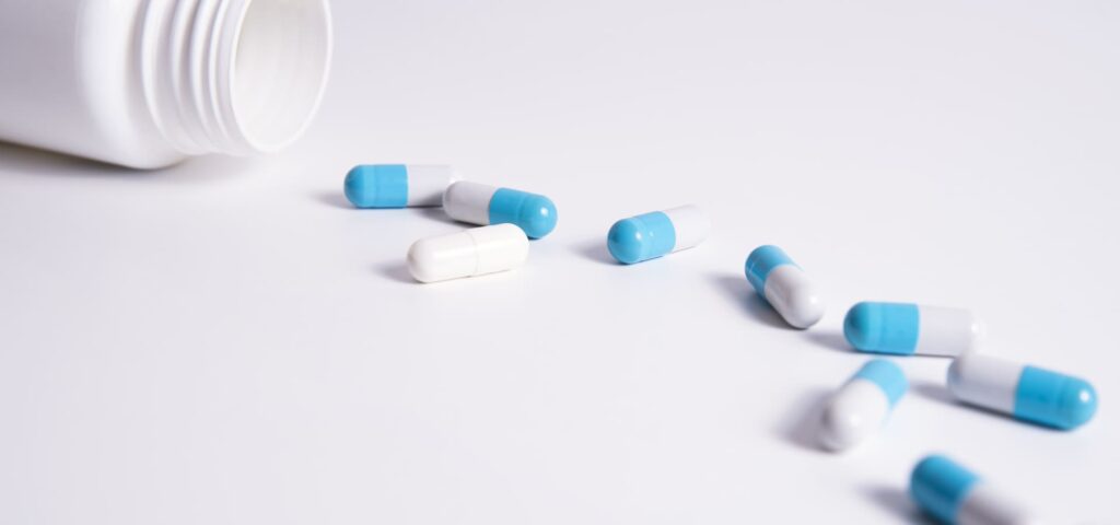 Capsules bleues et blanches étalées sur une surface blanche avec un flacon ouvert en arrière-plan.