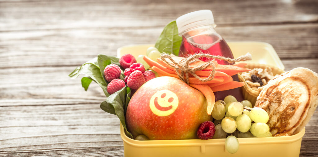 Un panier contenant une pomme décorée d'un visage souriant, des légumes frais (carottes, raisins secs, framboises), un jus et une pâtisserie.